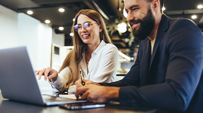 The Latest in Insurance Technology Adoption A woman in business attire smiling and pointing at an open laptop screen. A man in business attire writing down notes while listening to the woman. They are both sitting at a desk with the laptop in front of them