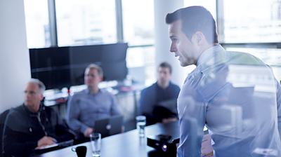 Merchants Insurance Group Automates Data Exchange with a Single Platform Man in business attire giving a presentation to 3 men sitting in boardroom with laptops and tablets in their hands